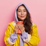 Pleased millennial girl with Asian appearance, has no makeup, wears violet sweatshirt and raincoat, holds flask with hot beverage, tries to warm while drinking tea, feels relaxed during rainy day