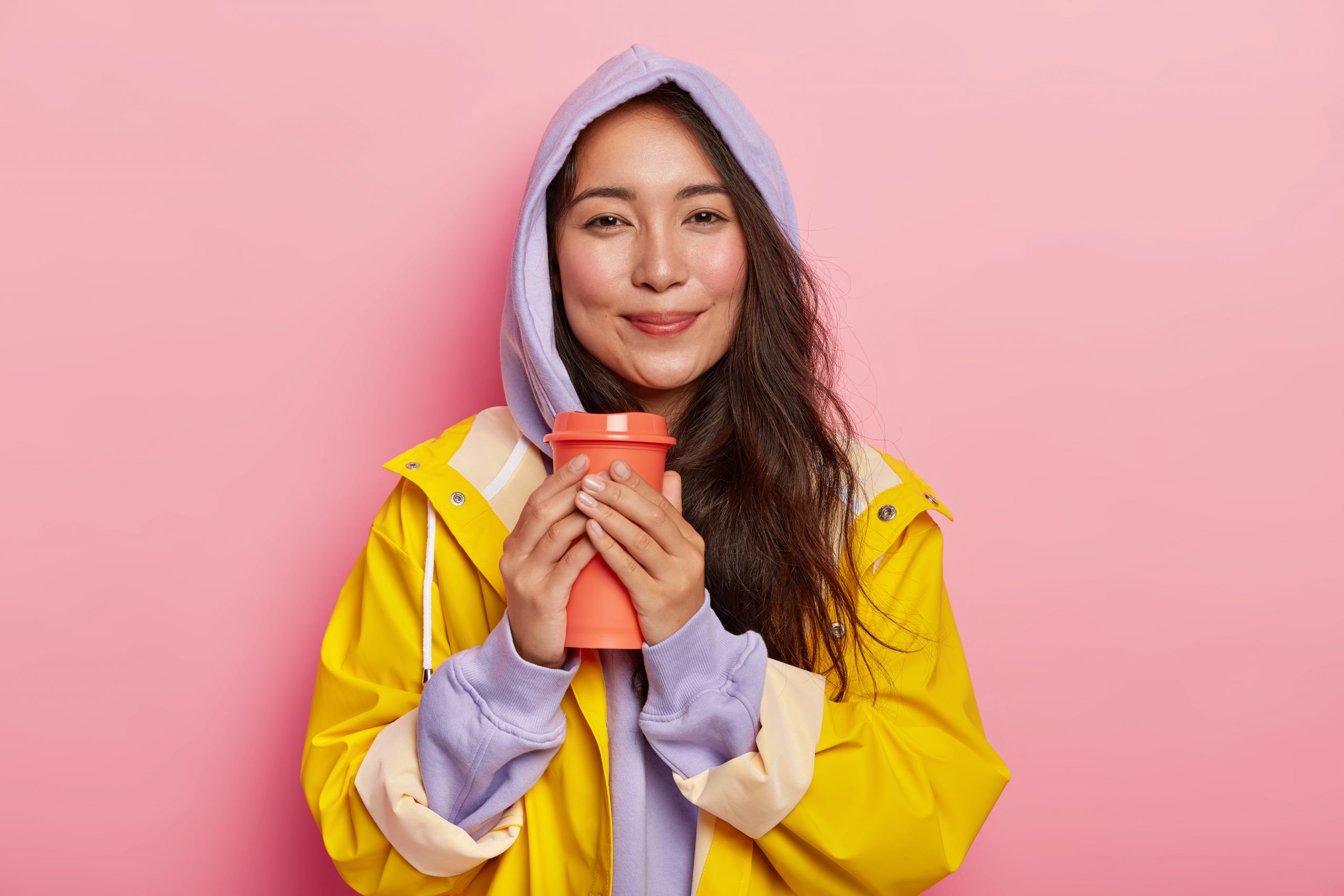 Pleased millennial girl with Asian appearance, has no makeup, wears violet sweatshirt and raincoat, holds flask with hot beverage, tries to warm while drinking tea, feels relaxed during rainy day