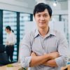 Portrait of successful handsome executive businessman smart casual wear looking at camera and smiling, arms crossed in modern office workplace. Young Asia guy standing in contemporary meeting room.