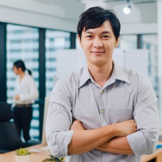 Portrait of successful handsome executive businessman smart casual wear looking at camera and smiling, arms crossed in modern office workplace. Young Asia guy standing in contemporary meeting room.