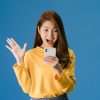 Surprised young Asia lady using mobile phone with positive expression, smiles broadly, dressed in casual clothing and standing isolated on blue background. Happy adorable glad woman rejoices success.